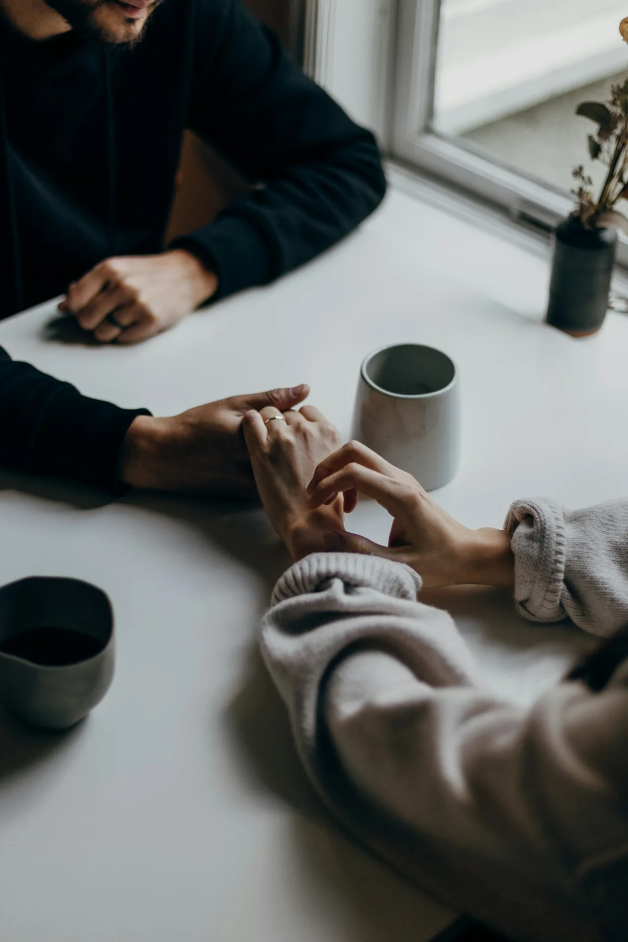 Two people hold hands across a light table beside ceramic mugs and a window.