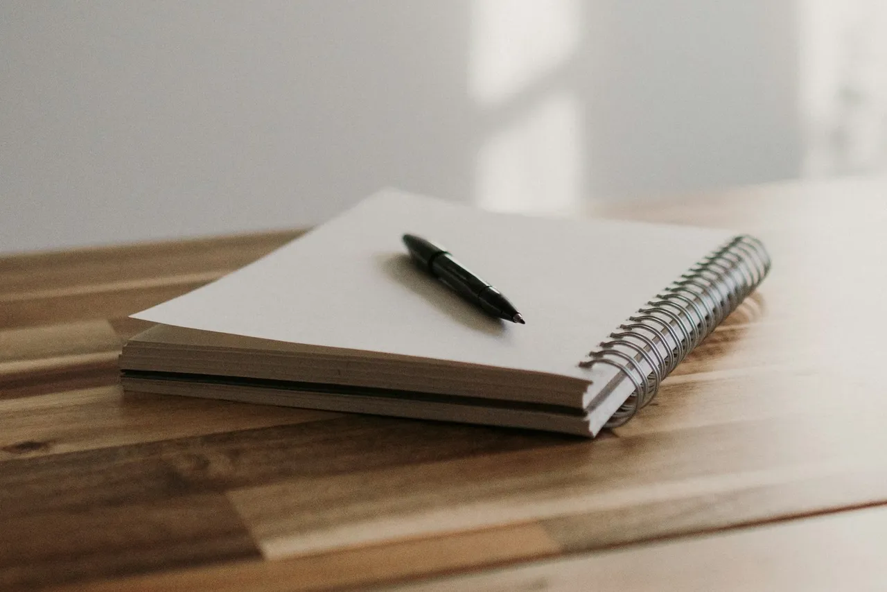 A blank spiral notebook and black pen sit on a wooden table beside soft window shadows.